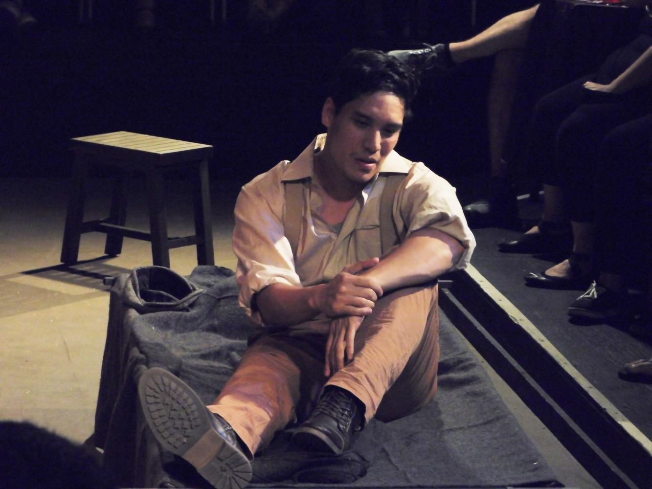 George (Nick Ono) talking about his friend Lennie on The Empty Space Theatre stage while sitting down and holding his left wrist.