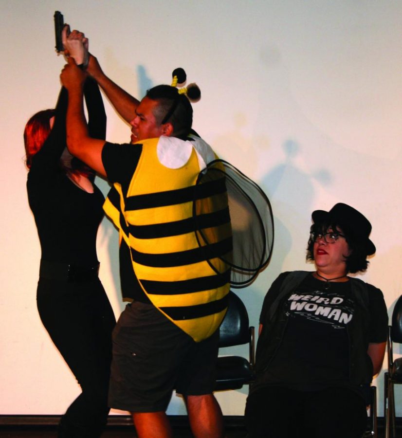 Claire Rock as Black Widow and Jesse Arenas as El Bumblo, struggle for control of the gun, while Alissa Morrow as DJ, looks on in dismay.