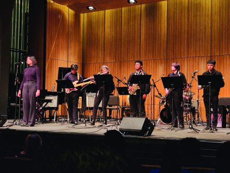 six musicians stand on stage with singer on far left and instrumentalists to her right