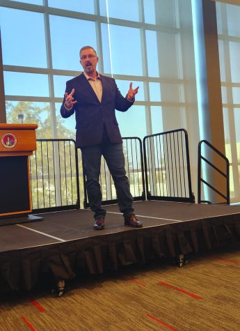 man in suit stands on stage wearing suit with his arms partially raise (bent at elbow) as he speaks to audience